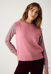 Pink ärmelloser Pullover mit strukturierter Oberfläche, getragen über einem gemusterten Langarmshirt mit roten und schwarzen Tropfenformen.