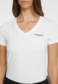 Weißes V-Ausschnitt-T-Shirt aus Baumwolle, mit einem kleinen mehrfarbigen 'GUESS JEANS'-Logo auf der linken Brustseite, kurzen Ärmeln und einer figurbetonten Passform.