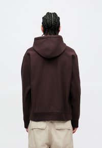 Felpa marrone con vestibilità comoda, dotata di cappuccio con coulisse, polsini a costine e una texture morbida. Indossata con pantaloni beige chiaro, vista da dietro.