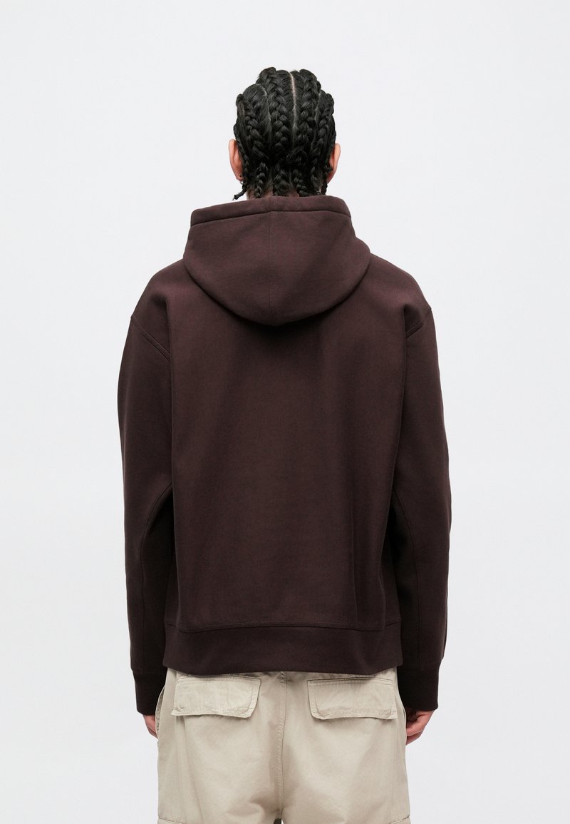 Brun hættetrøje med løs pasform, der har justerbar hætte med snøre, ribbet kant ved ærmerne og blød tekstur. Bær den sammen med lysebeige bukser, set bagfra.