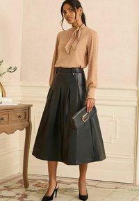 Bézs kötött blúz nyakán masnival, fekete pliszírozott műbőr midi szoknyával és fekete magassarkú cipőt viselve, kezében egy clutch-t tartva.