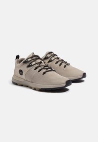 Zapatillas de malla beige con cordones negros, suelas de goma y detalle del logo. Presenta una parte superior texturizada y un soporte robusto en el talón para mayor comodidad.