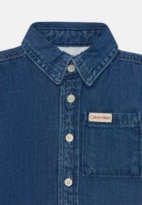 Tamsiai mėlynas džinsinis marškiniai su mygtukų uždarymu, smailiu apykakle ir krūtinės kišene, ant kurios yra mažas Calvin Klein ženklas. Lygi tekstūra.