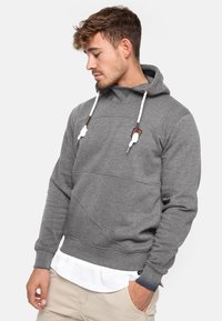 Grauer Kapuzenpullover mit strukturiertem Finish, ausgestattet mit einer Kängurutasche, weißen Kordeln und kontrastierenden braunen Akzenten an der Kapuze.