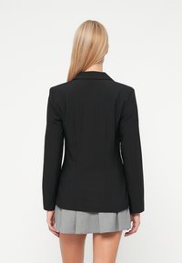 Μαύρο προσαρμοσμένο σακάκι με πέτο και μακριά μανίκια, με κολακευτική σιλουέτα. Το μοντέλο φορά μια γκρι πλισέ φούστα.