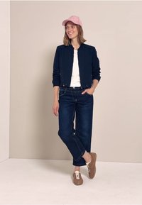 Chaqueta bomber azul marino, camiseta blanca, jeans azul oscuro y zapatillas marrones. Modelo lleva una gorra rosa clara, de pie contra un fondo neutro.