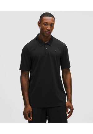 Mees seisab ühevärvilise taustaga ees, kandes musta lühikeste varrukatega polosärki, millel on rinnal väike logo.