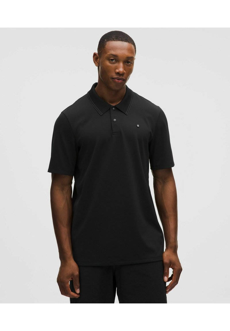 Mężczyzna stojący na jednolitym tle, ubrany w czarną koszulkę polo z krótkim rękawem i małym logo na klatce piersiowej.