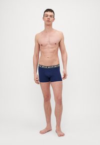 Jūras zila vīriešu bokseršortos ar melnu zīmola jostu, pieguļošs dizains un gluda tekstūra. Tos valkā modelis, stāvot basām kājām.