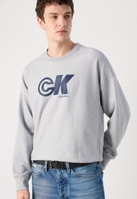 Šedá mikina vyrobená z bavlněné směsi, s velkým námořnickým logem "CK" a textem "Calvin Klein". Dlouhé rukávy s uvolněným střihem a kulatým výstřihem.
