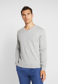Szary sweter typu V-neck z długimi rękawami, ściągaczami przy rękawach i dołem. Tkanina wydaje się miękka z delikatną teksturą. Małe logo na klatce piersiowej.