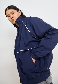 Mørkeblå vindtæt jakke med oversized pasform, hvid kantbånd, elastiske manchetter, høj krave og sidetasker. Blødt, let materiale.