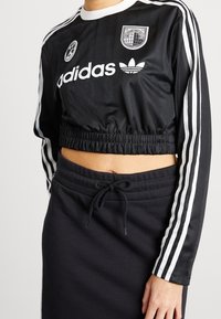Person som bär en svart Adidas kortärmad långärmad topp med vita ränder och logotyper, i kombination med svarta dragsko byxor eller kjol.