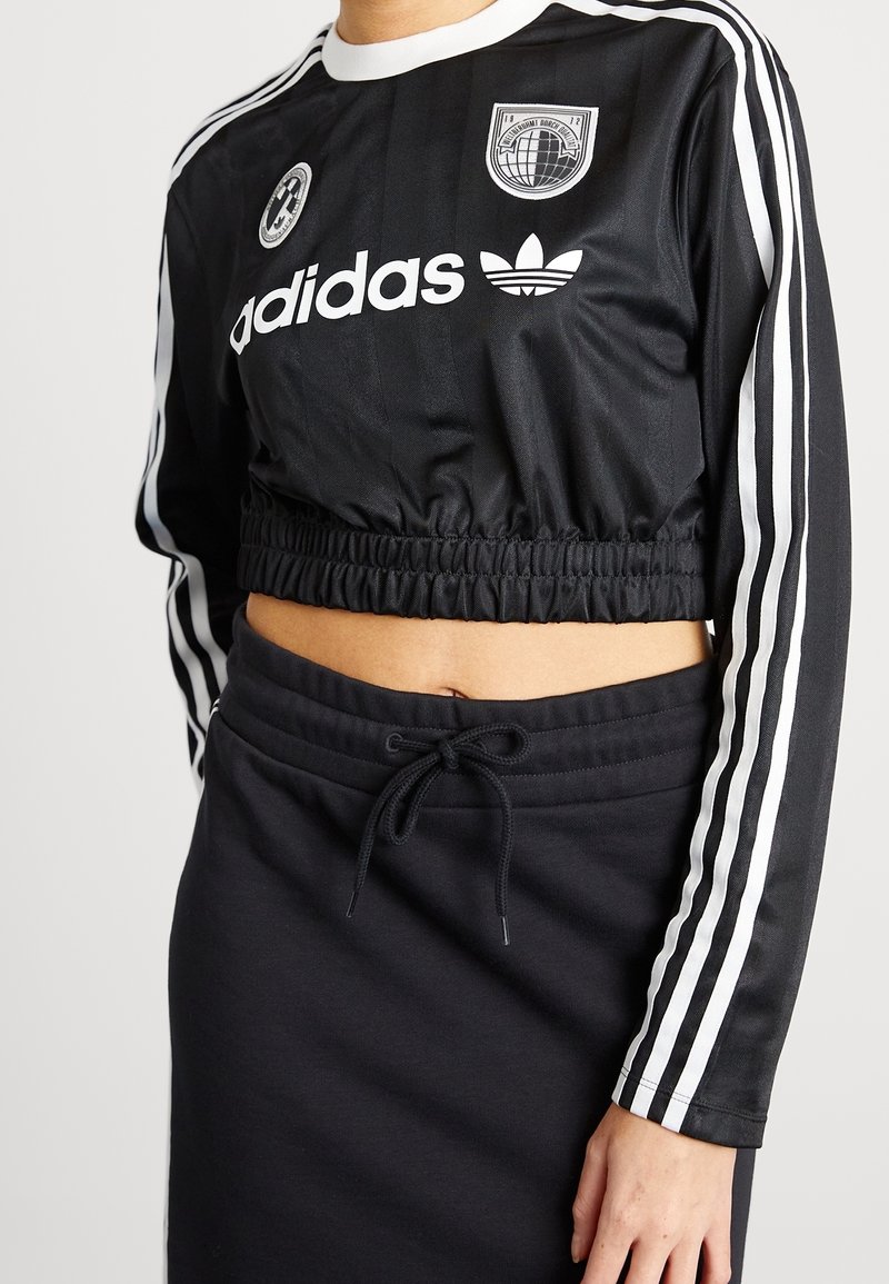 Person som bär en svart Adidas kortärmad långärmad topp med vita ränder och logotyper, i kombination med svarta dragsko byxor eller kjol.