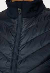 Laivastonsininen toppatakki korkealla kauluksella, jossa on etuvetoketju ja viistokuvioitu tikkaus. Sisältää kiiltävän logodetaillin kauluksessa.