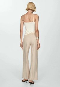 Débardeur en crème à fines bretelles associé à un pantalon beige taille haute et large. Le mannequin se tient devant un fond blanc uni.