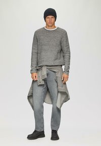Grijze gebreide trui met een ronde hals, lichtgrijze shirt om de taille geknoopt, grijze jeans en zwarte schoenen. Model draagt een donkere beanie.