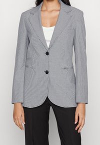 Šedý trenčkot ve vzoru houndstooth s dvěma černými knoflíky, klopy s výřezy a dvěma předními kapsami. Disponuje dlouhými rukávy a přiléhavým střihem.