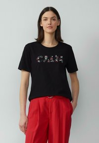 Zwarte katoenen t-shirt met korte mouwen, voorzien van een kleurrijk versierd tekstontwerp aan de voorkant. Gecombineerd met rode geplooide broek.
