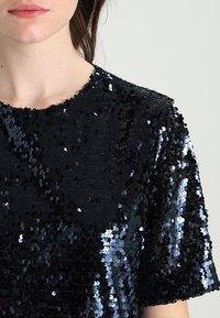 Svart paljett-topp med rund halsringning och korta ärmar. Tyget har en glänsande, strukturerad yta som skapats av överlappande paljetter.