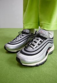 Stříbrné tenisky Nike Air Max 97 mají černé akcenty, síťovaný svršek s vlnitými designovými liniemi a růžové logo na boku.