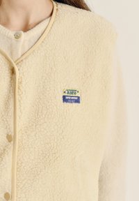 Kremo spalvos vilnos liemenė su smėlio spalvos apvadu, apvali iškirptė, spaudžiami mygtukai ir mėlynos bei žalios spalvos etikete su užrašu „AMV SUPER VINTAGE".