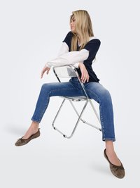 Jeune femme aux longs cheveux blonds portant un gilet bleu marine, une chemise blanche, un jean et des ballerines marron, assise à l'envers sur une chaise pliante en métal.