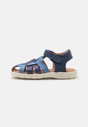 Blå och marinblå sandal med korsade läderremmar, öppen tåsdesign, justerbar vristrem och en ljusfärgad gummisula.