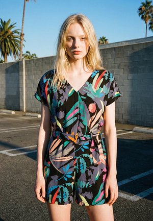 Jeune femme aux cheveux blonds se tenant dans un parking ensoleillé, portant une combinaison courte à manches courtes avec ceinture et motif floral coloré.
