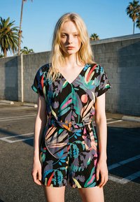 Tânără cu păr blond stă într-o parcare însorită, purtând un costum scurt cu mâneci scurte, cu model floral colorat și curea.