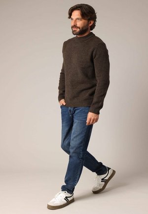 Pull en maille marron avec un col montant, associé à un jean bleu et des baskets blanches avec des accents noirs et des semelles grises. Modèle se tenant de côté.