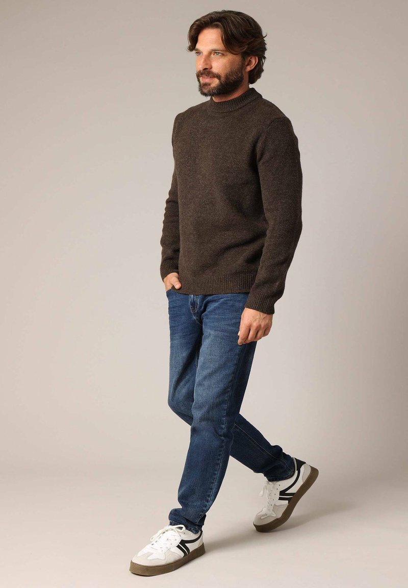 Bruine gebreide trui met een hoge hals, gecombineerd met blauwe jeans en witte sneakers met zwarte accenten en grijze zolen. Model staat zijwaarts.