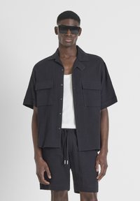 Zwarte kortmelding knoopshirt met twee borstzakken, gecombineerd met bijpassende shorts. Witte tanktop eronder, met een gladde stoftextuur.