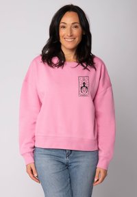 Lichtroze sweatshirt gemaakt van zachte stof, met een zwart grafisch ontwerp op de borst met elementen van een leeuw en het woord "STERKTE."