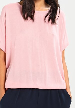 Femme portant un chemisier ample rose clair à manches courtes et un pantalon bleu marine, mains dans les poches, sur un fond uni.