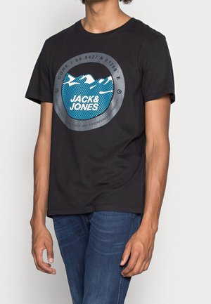 Mężczyzna ubrany w czarny T-shirt z niebiesko-białym okrągłym logo "Jack & Jones", zestawiony z ciemnoniebieskimi dżinsami, stojący na tle białej ściany.