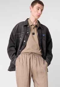 Veste en denim noir à coupe décontractée, fermeture à boutons et étiquette bleue. Polo beige avec étiquette et pantalon à cordon de serrage beige clair, texture lisse.