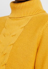 Gestrickter gelber Pullover mit hochgezogenem, geripptem Kragen, der ein Zopfmuster auf der Vorderseite sowie eine strukturierte, weiche Oberfläche aufweist.