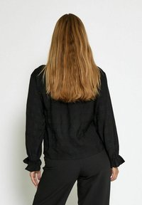 Blusa nera a maniche lunghe con tessuto testurizzato, polsini arricciati e vestibilità morbida, abbinata a pantaloni neri, vista da dietro.