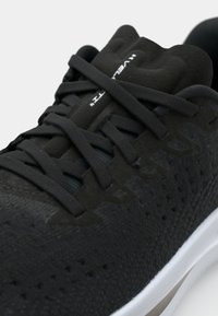 Zapato deportivo negro con parte superior texturizada, puntera redondeada, cordones planos y entresuela blanca con un sutil detalle beige.