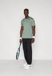 Grønn T-skjorte med korte ermer og "BOSS" logo, sammen med svarte joggebukser, hvite tennissko og holder en blå og svart tenniskjøtt.