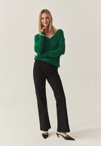 Grüner Strick-V-Ausschnitt-Pullover, schwarze lockere Jeans und schwarze spitze Schuhe. Glatte Textur, lässige Passform, minimales sichtbares Zubehör.