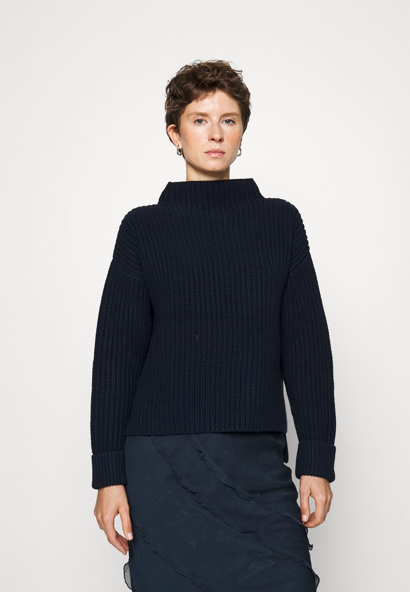 Giovane con capelli corti e ricci, che indossa un maglione a coste blu navy e una gonna nera a balze, in piedi davanti a uno sfondo bianco uniforme.