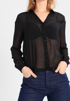 Frau trägt eine schwarze durchsichtige gepunktete Bluse mit Knöpfen über einem schwarzen BH, kombiniert mit dunkelblauen Jeans, Hände in den Taschen.