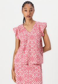 V-Ausschnitt-Oberteil aus leichtem Stoff mit einem rosa geometrischen Muster und Rüschensäumen. Ein zusätzlicher Rock im passenden Design ist sichtbar.