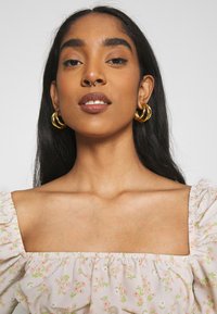 Top rosa floral de ombros à mostra, com mangas bufantes gathered, decote elástico e pequenos padrões de flores verdes e rosas. Brincos dourados em forma de argola.