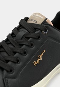 Schwarzer Sneaker aus strukturiertem Leder mit kontrastierender weißer Sohle, braunen Akzenten, schwarzen Schnürsenkeln und einem goldenen Logo an der Seite.