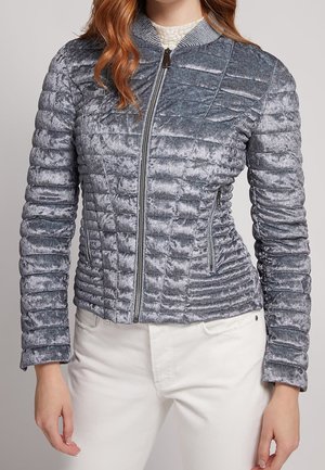 Vrouw draagt een aansluitend, grijs, gewatteerd fluwelen jasje met een ritssluiting aan de voorkant en een gestreepte kraag, gecombineerd met een witte broek.