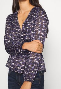 Lila gemusterte Bluse mit V-Ausschnitt, langen Ärmeln und Raffung an der Taille. Hergestellt aus leichtem Stoff mit fließender Textur.