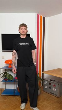 Νεαρός άνδρας με τατουάζ, ντυμένος με μαύρο μπλουζάκι και παντελόνι Ripcurl, στέκεται σε ένα δωμάτιο με τηλεόραση, πορτοκαλί φωτιστικό και ριγέ διακόσμηση στον τοίχο.